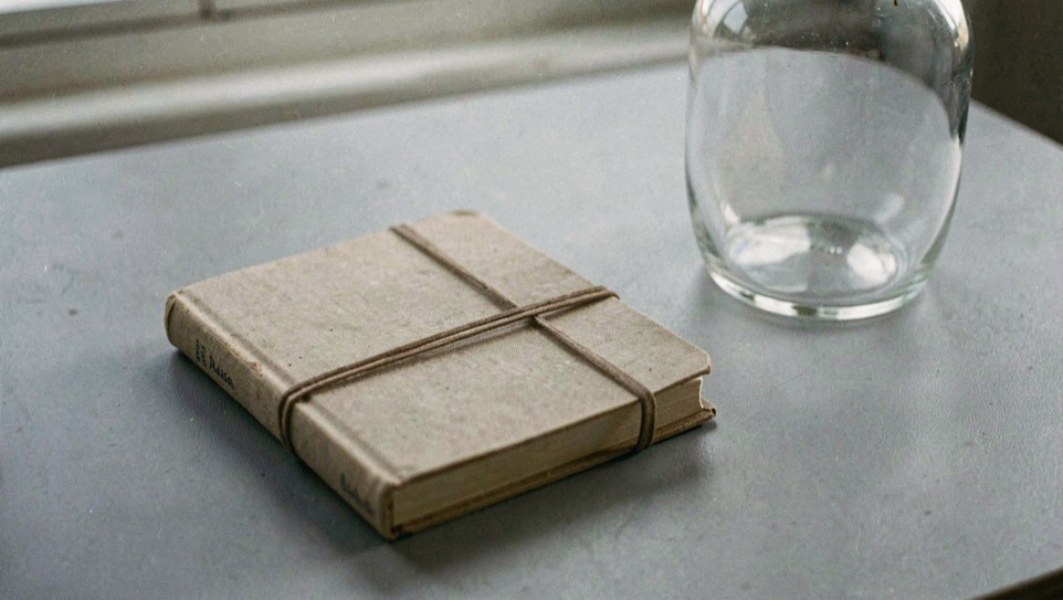 Gesloten notitieboek met een elastiek eromheen ligt op een egaal grijs bureaublad naast een lege glazen vaas